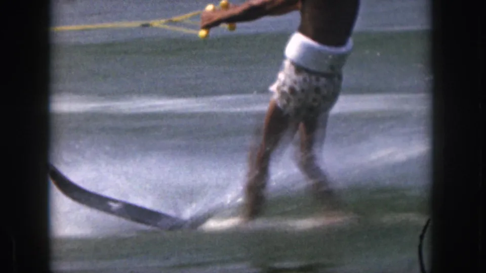 In the 1961 Michigan home movie, a shirtless adult man in patterned swim trunks holds a yellow tow rope while skiing across lake water.