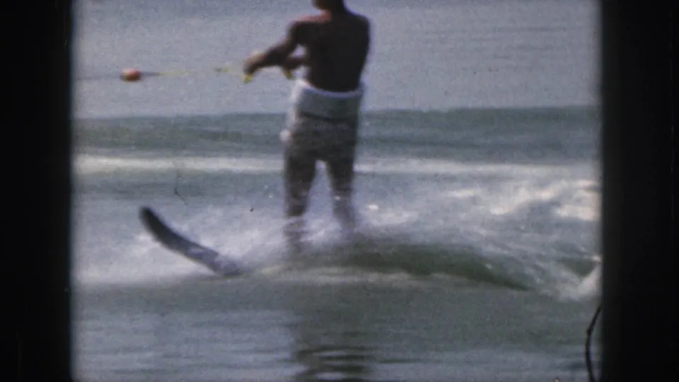 A rear view shows the adult skier moving across the Michigan lake, holding the tow rope as a small ski and spray appear behind him.