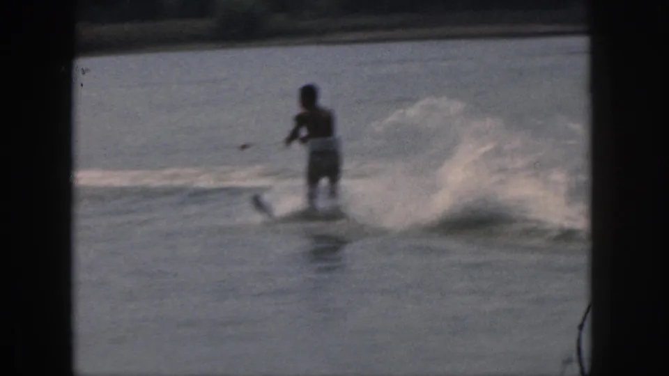 1961 Michigan home-movie frame of a man water skiing away from the camera, with a low wake and soft film grain.