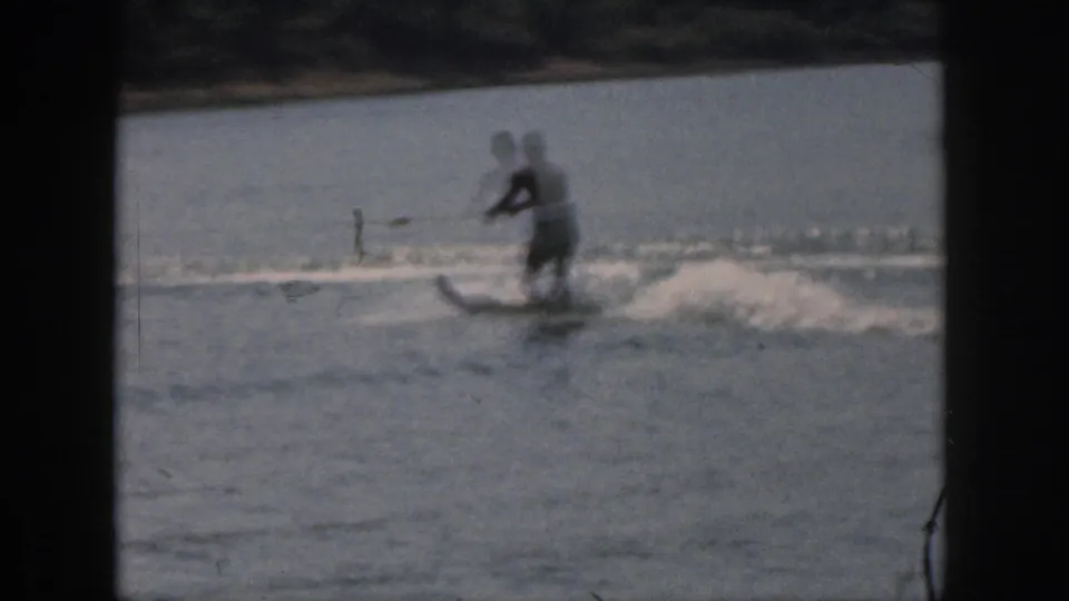 Soft 8mm frame from Michigan in 1961 showing a man on water skis, tow rope, lake wake, and distant shoreline.