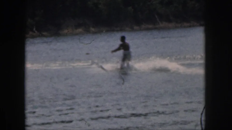 1961 Michigan 8mm frame showing a man water skiing near a tree-lined shore, with white spray and visible film wear.