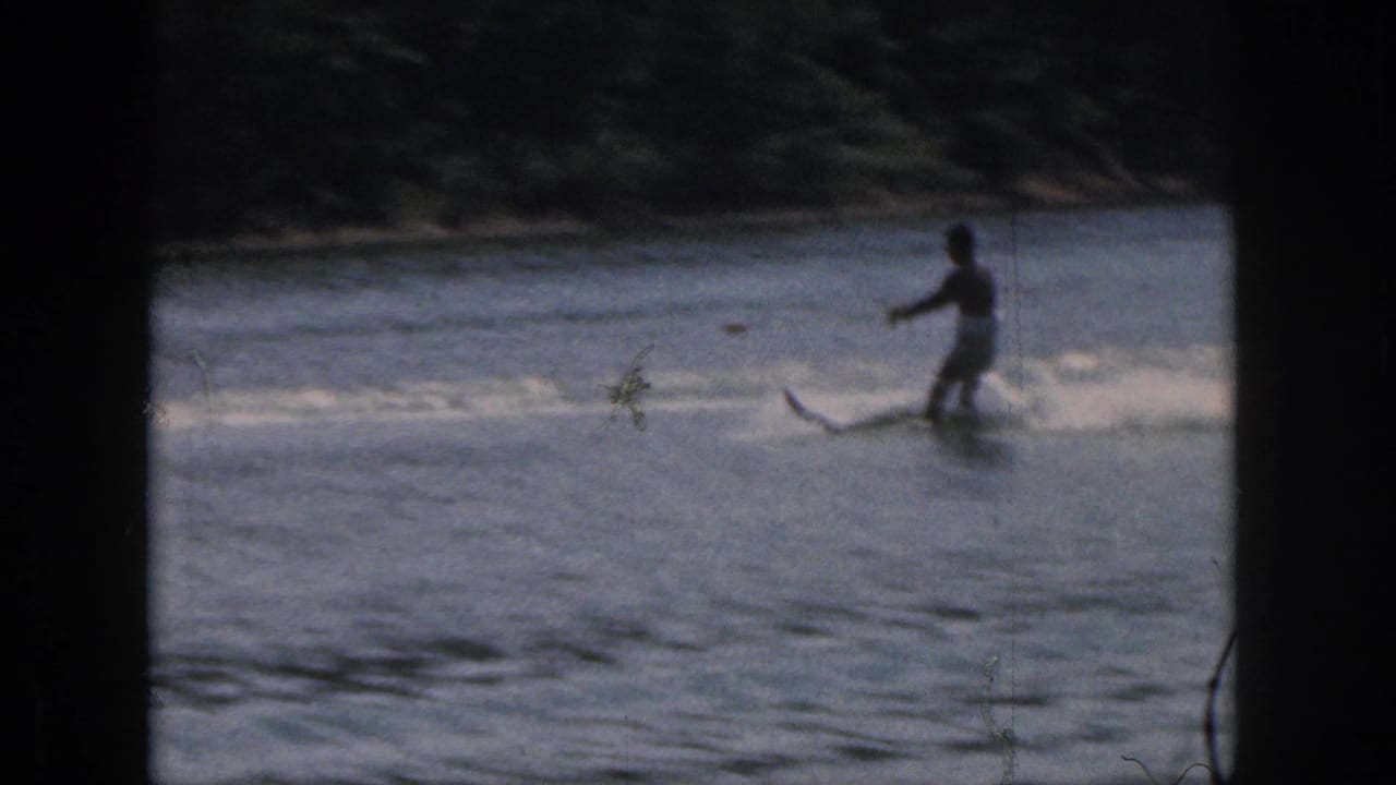 1961 Michigan 8mm frame showing a distant water skier crossing lake water near a dark wooded shoreline.