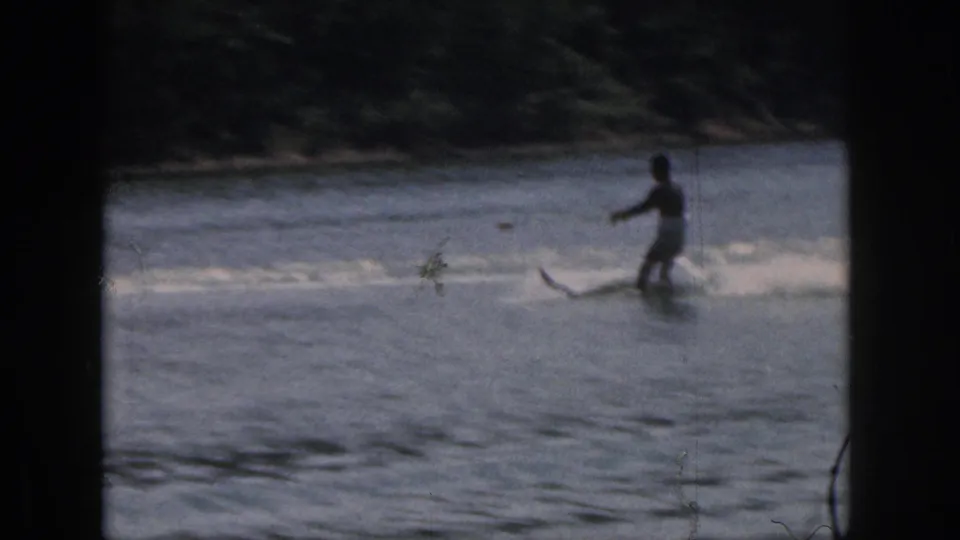 1961 Michigan 8mm frame showing a distant water skier crossing lake water near a dark wooded shoreline.