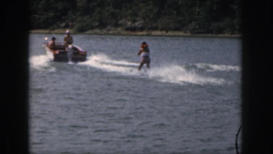 1961 Michigan home-movie frame with a skier being towed behind a motorboat near a wooded lake shoreline.