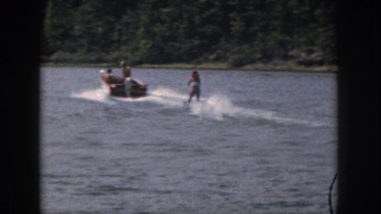 Woman water skier trails a motorboat across open lake water near a wooded shore.