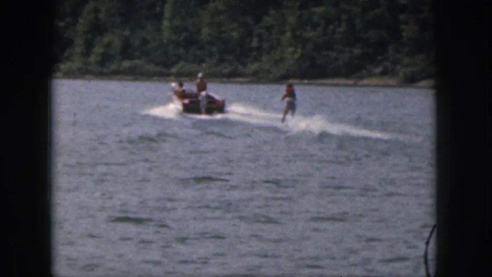 1961 Michigan 8mm still with a motorboat and woman water skier moving across a wooded lake scene.