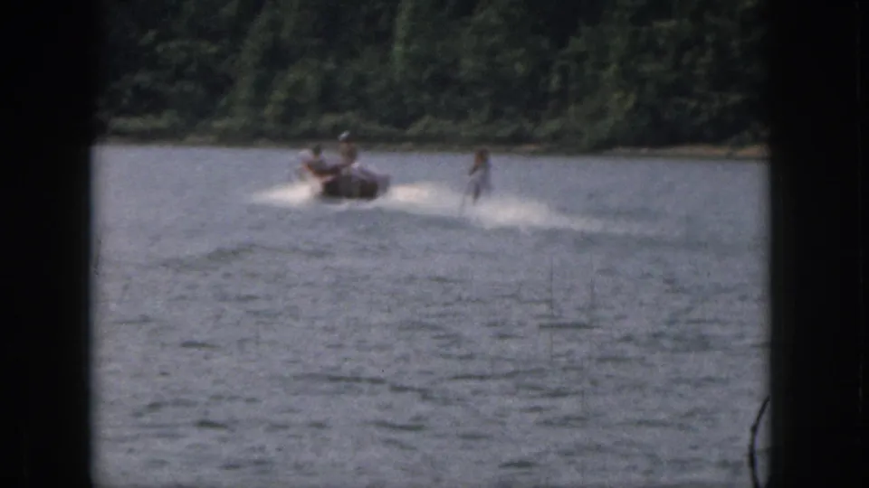 1961 Michigan home-movie frame showing a distant boat towing a woman water skier through lake spray.