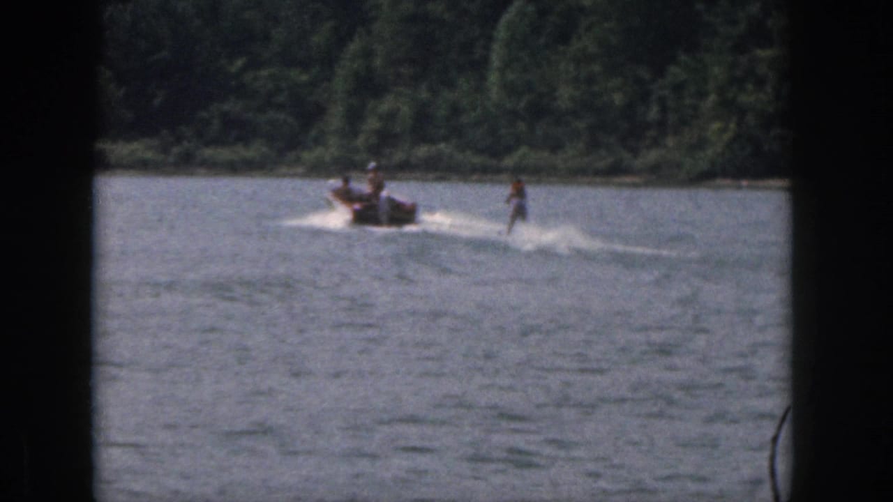 1961 Michigan 8mm frame showing a distant motorboat towing a water skier across a sunny lake with trees along the shore.