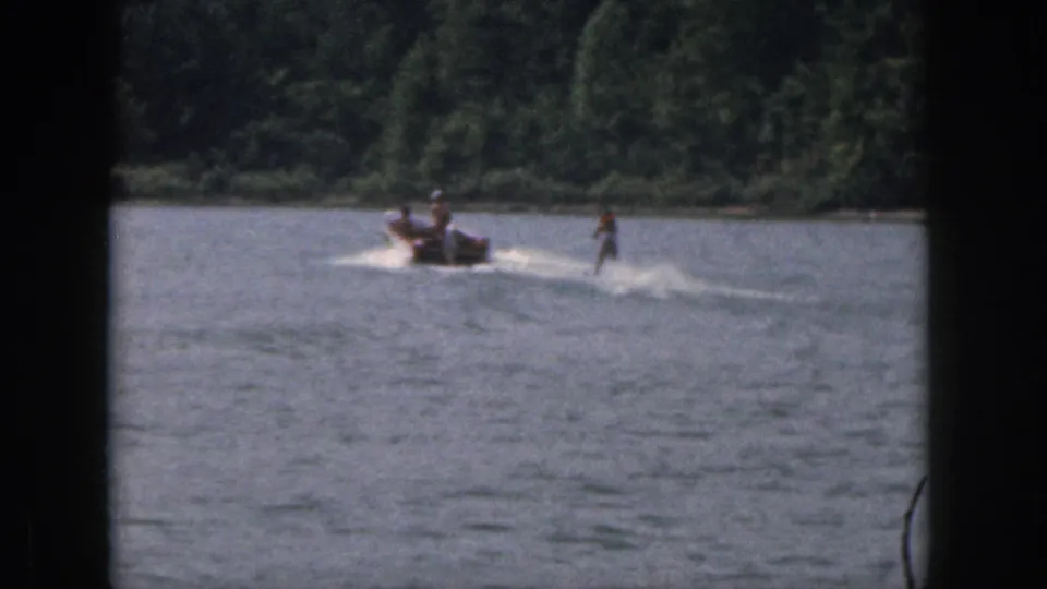 1961 Michigan 8mm frame showing a distant motorboat towing a water skier across a sunny lake with trees along the shore.