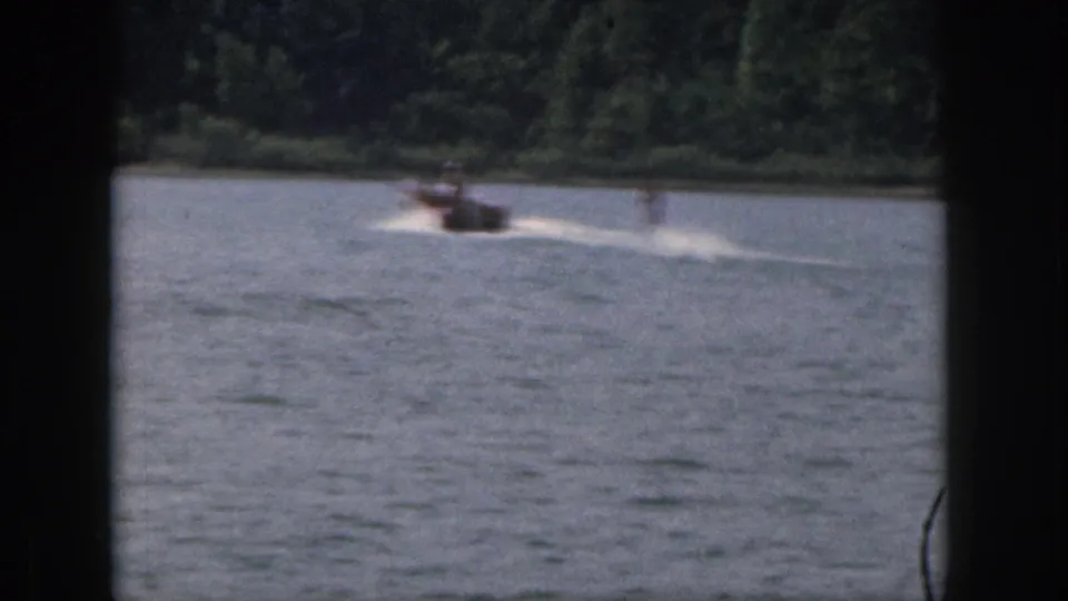 Distant 1961 Michigan lake view with a motorboat crossing the frame and the skier less distinct in the wake.