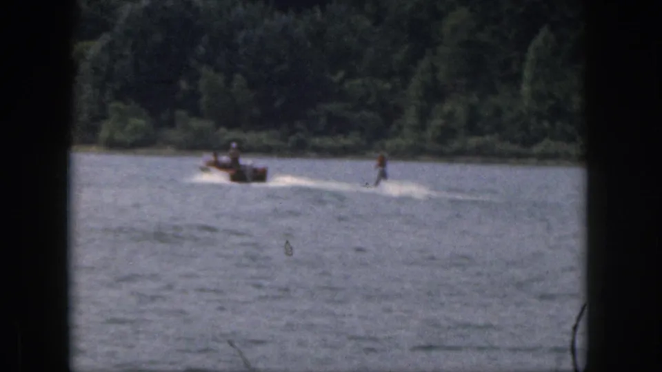 1961 Michigan home-movie frame of a skier holding position behind a motorboat on a calm lake bordered by trees.