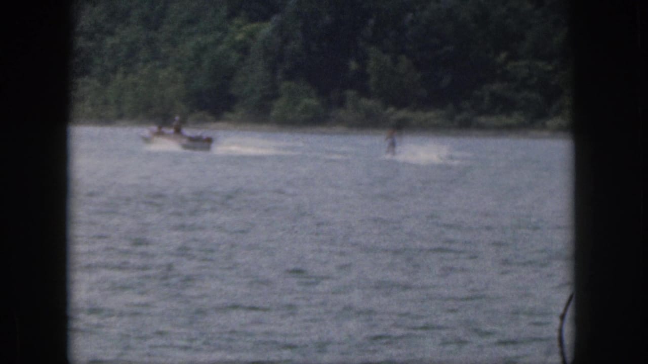 1961 Michigan home-movie frame showing a small boat pulling a distant water skier across a tree-lined lake.