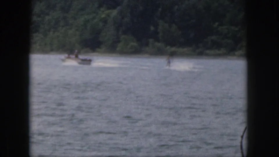 1961 Michigan home-movie frame showing a small boat pulling a distant water skier across a tree-lined lake.