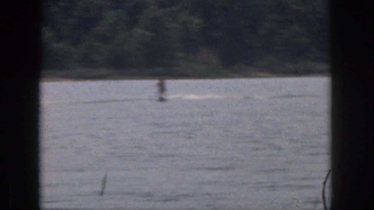 Distant water skier crossing a Michigan lake in 1961, framed by dark 8mm borders and a wooded bank.
