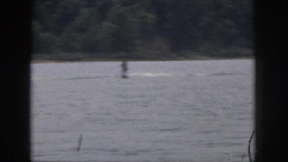 Distant water skier crossing a Michigan lake in 1961, framed by dark 8mm borders and a wooded bank.