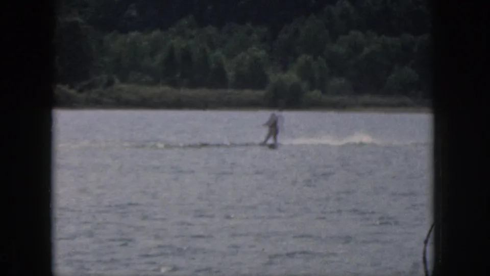 Distant 1961 Michigan water-skiing frame with the skier moving across a lake before a dense wooded shoreline.