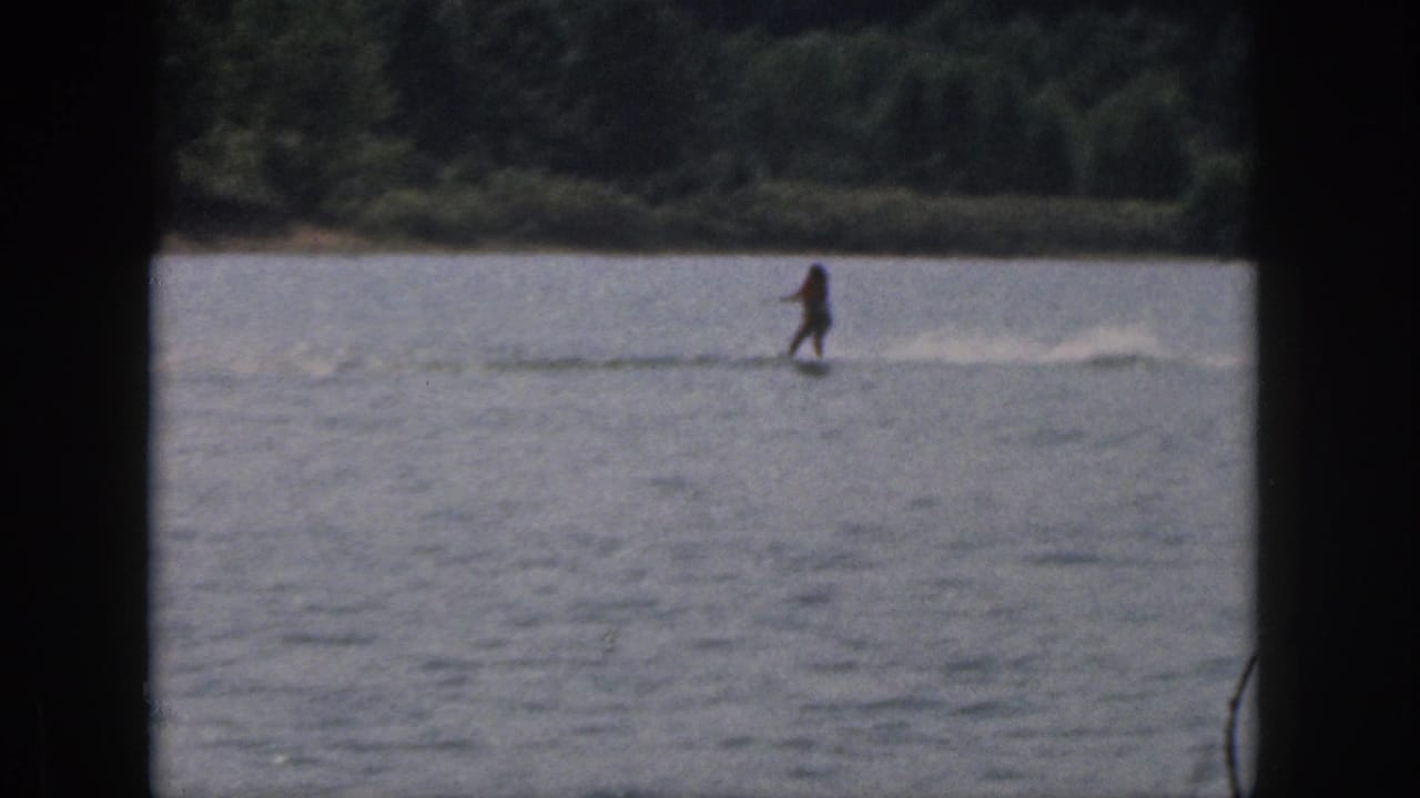 A distant water skier moves across a Michigan lake in 1961 home-movie footage, with a low wake and tree-lined shore in the background.