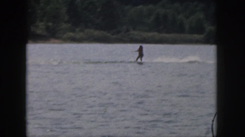 A distant water skier moves across a Michigan lake in 1961 home-movie footage, with a low wake and tree-lined shore in the background.