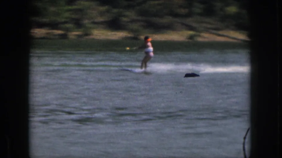 A woman in a light swimsuit water skis across a Michigan lake, leaving white spray with a wooded shoreline behind her.