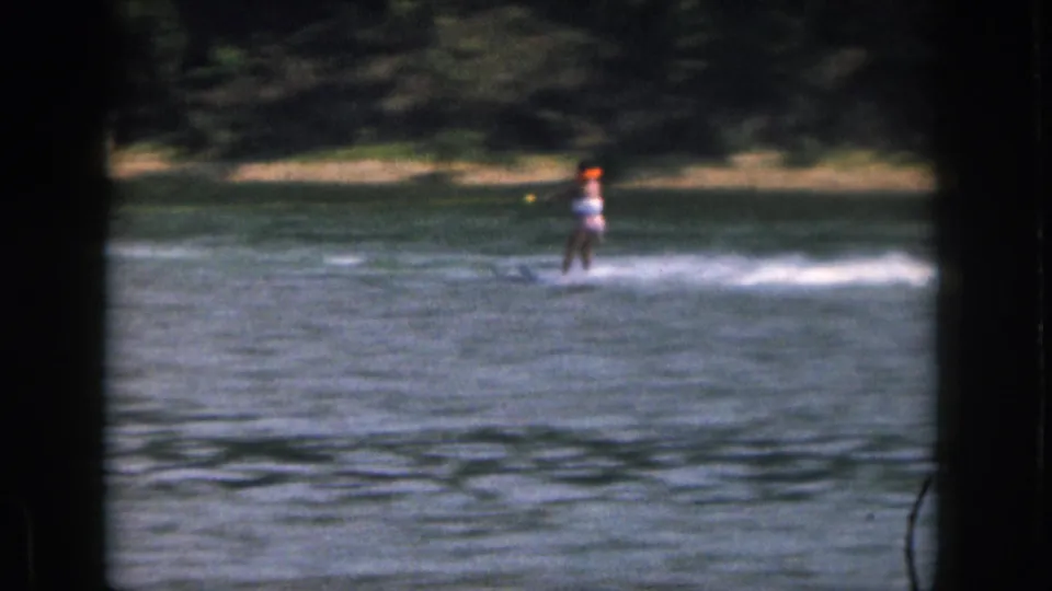 The skier continues across the lake in soft 8mm color, holding balance as spray stretches behind her near the tree-lined shore.