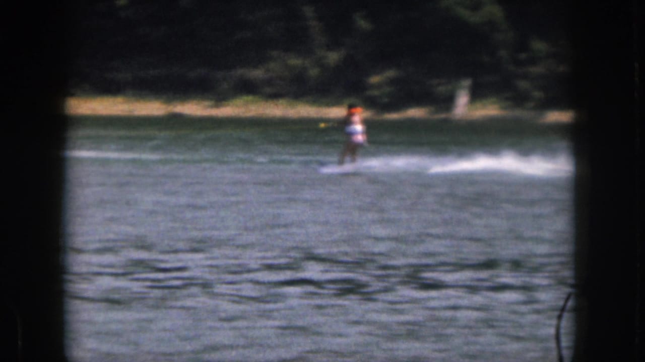 1961 Michigan 8mm frame showing a distant water skier in an orange life vest moving across a calm lake near trees.