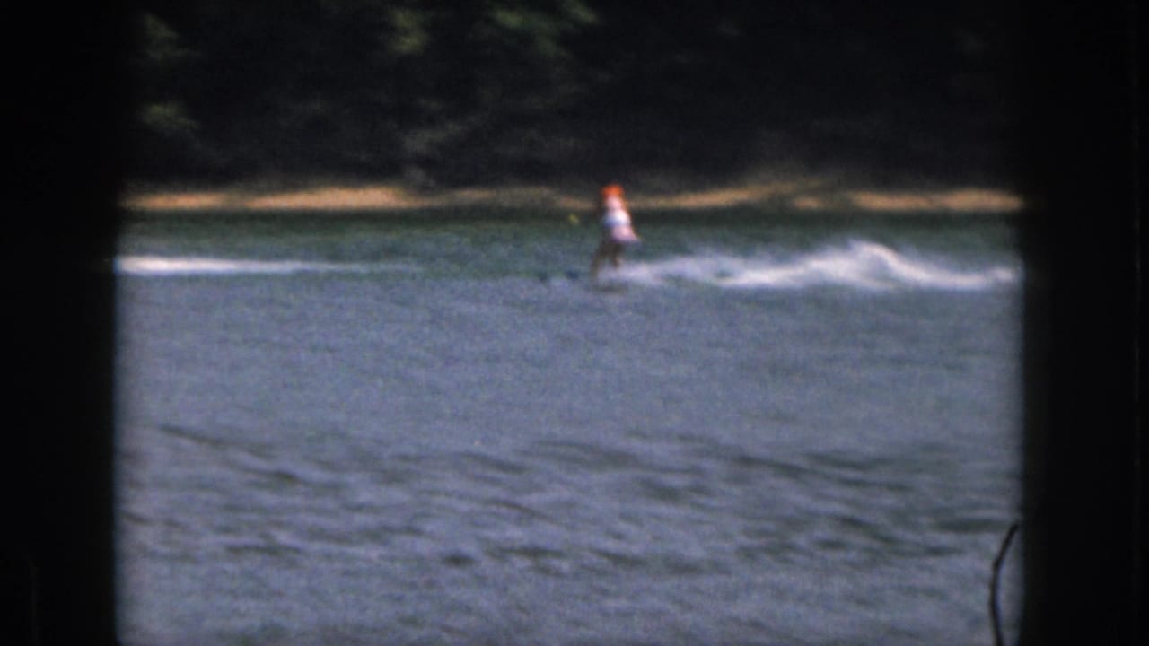 A distant skier in bright safety gear moves across the Michigan lake, framed by soft 8mm borders and shoreline trees.