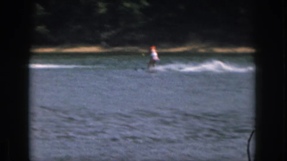 A distant skier in bright safety gear moves across the Michigan lake, framed by soft 8mm borders and shoreline trees.