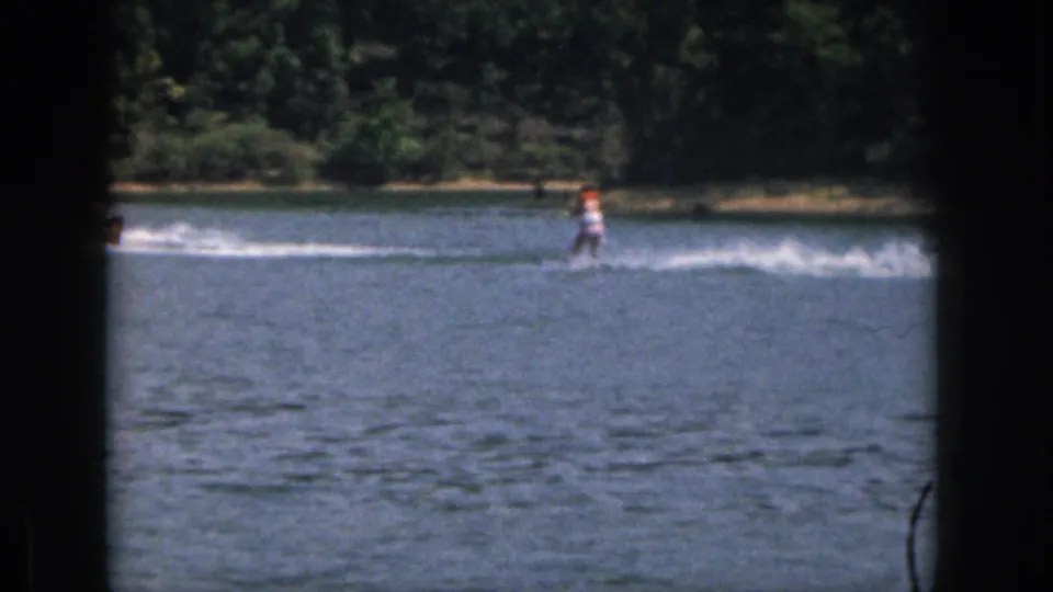 1961 home-movie frame of a water skier on a Michigan lake, with dense shoreline trees and soft color 8mm grain.
