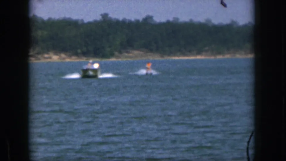 A small motorboat pulls a distant water skier across a Michigan lake in 1961, with shoreline trees and visible 8mm film borders.