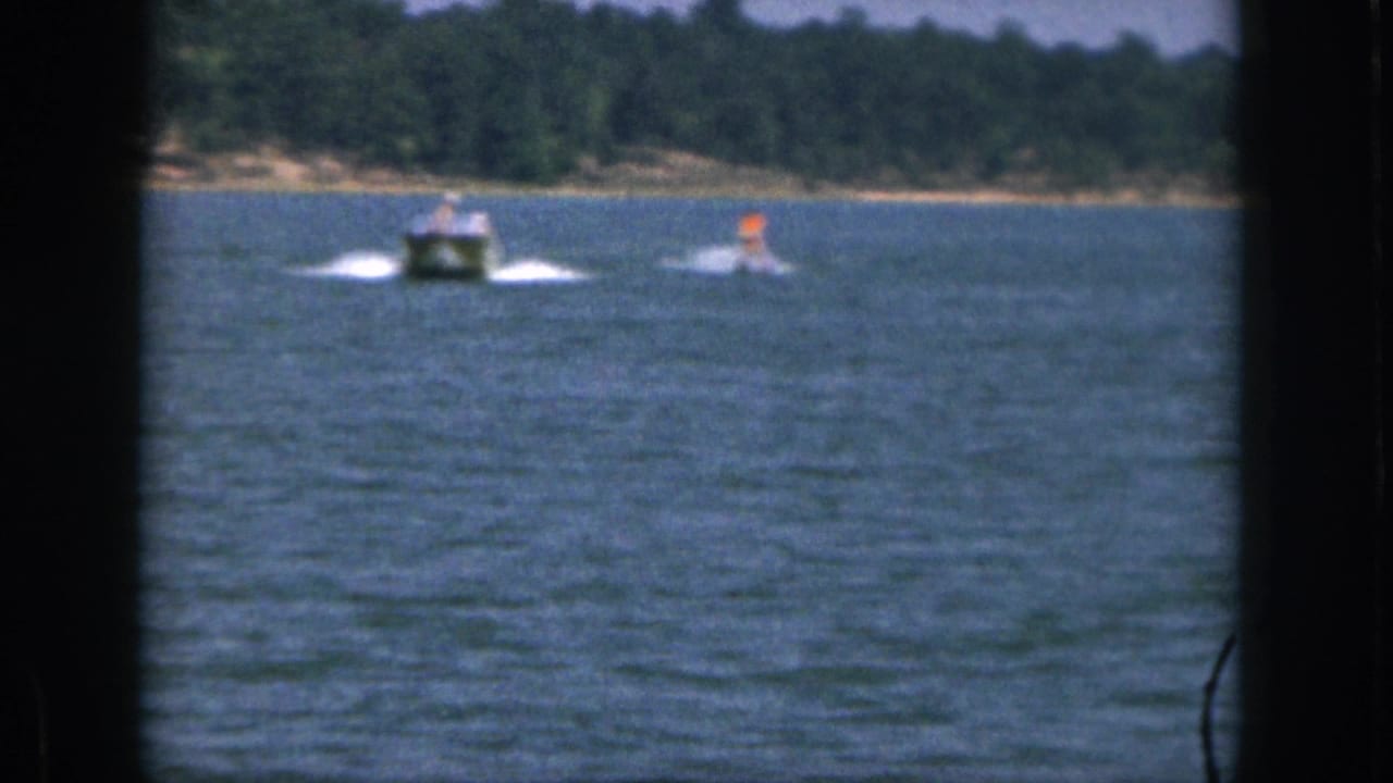 1961 Michigan 8mm frame showing a small motorboat towing a distant water skier in orange across blue lake water.
