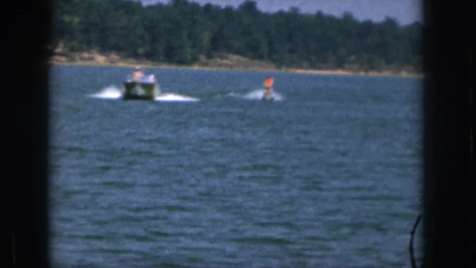 1961 Michigan home-movie frame with a motorboat and orange-clad skier moving over a lake before a wooded shore.