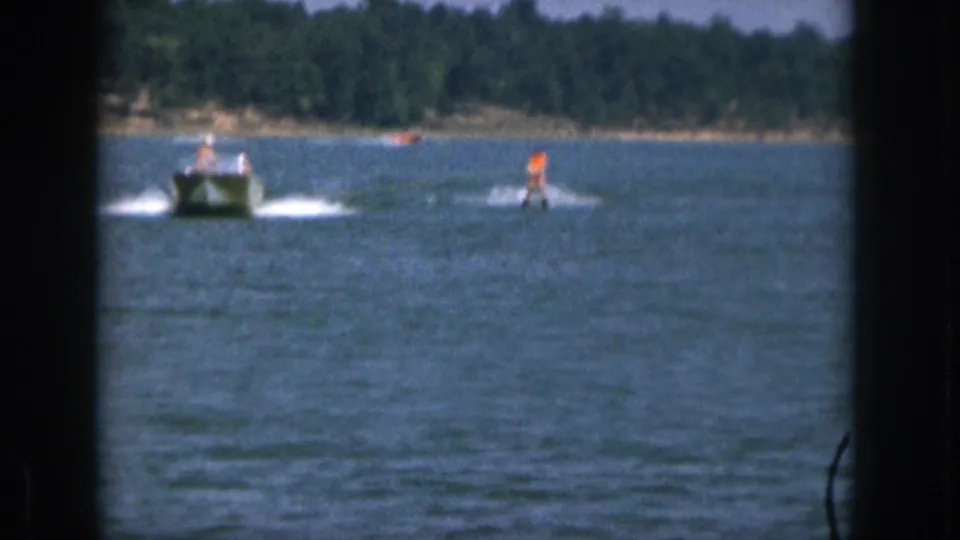 1961 Michigan home-movie frame with a distant orange-clad water skier following a small boat across blue lake water.