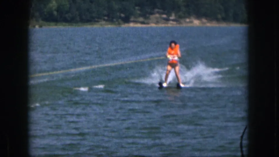 1961 Michigan home-movie frame of a waterskier in an orange life vest cutting across lake water near a wooded shore.