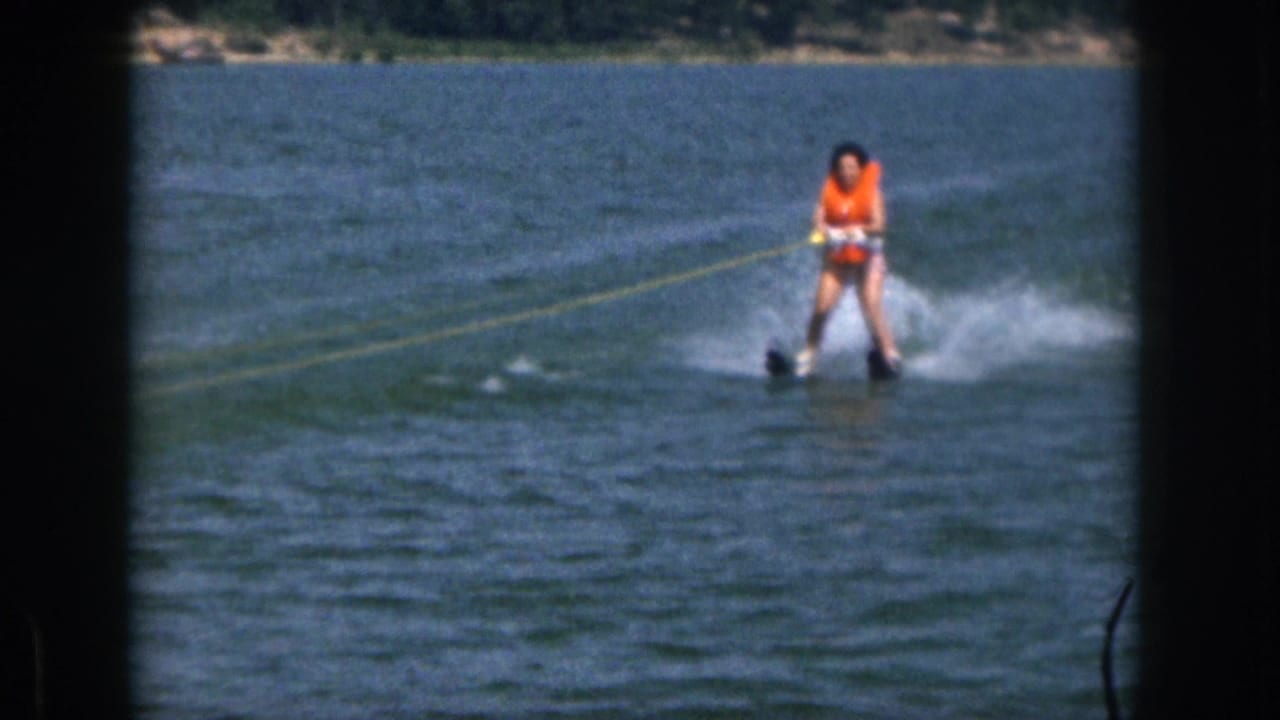 In 1961 Michigan home-movie footage, a woman in an orange life vest water skis across a lake with a wooded shoreline behind her.