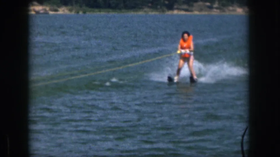 In 1961 Michigan home-movie footage, a woman in an orange life vest water skis across a lake with a wooded shoreline behind her.