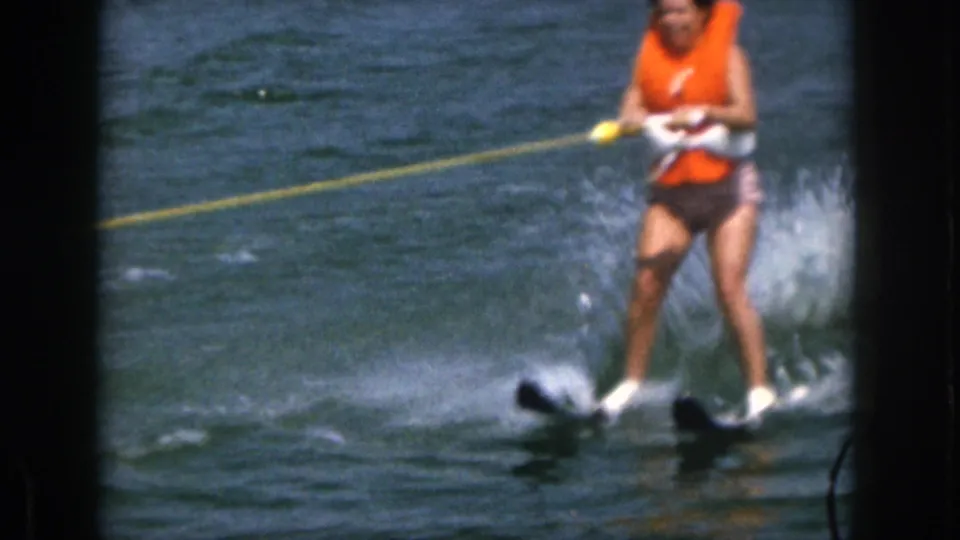 Closer 8mm view of the woman water skiing in an orange life vest, gripping the tow handle as lake spray rises around her skis.
