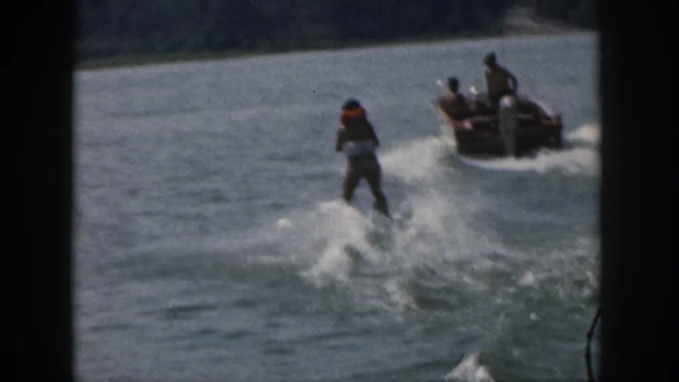 1961 Michigan home-movie frame with a skier riding the wake behind a motorboat on open lake water.