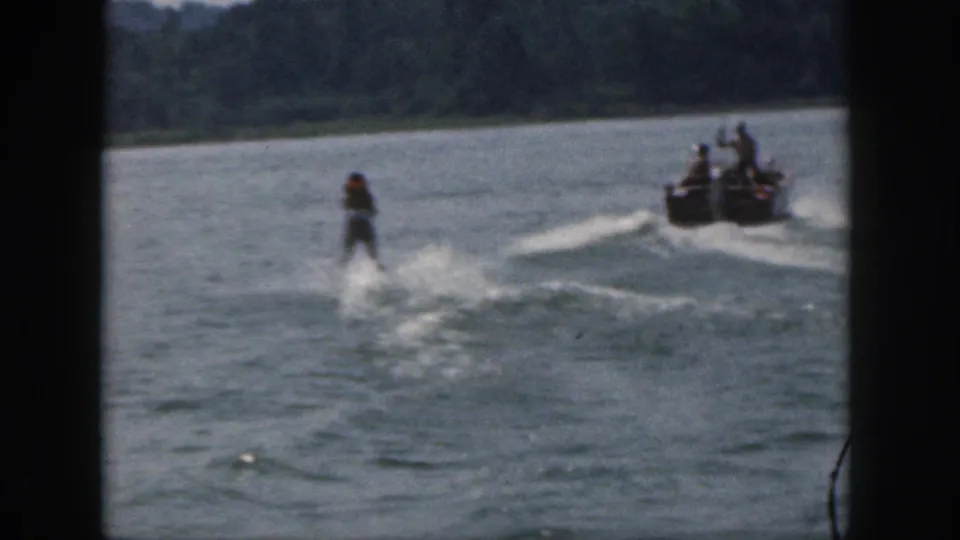 1961 Michigan home-movie frame with a skier and motorboat receding across the lake, framed by 8mm gate edges.