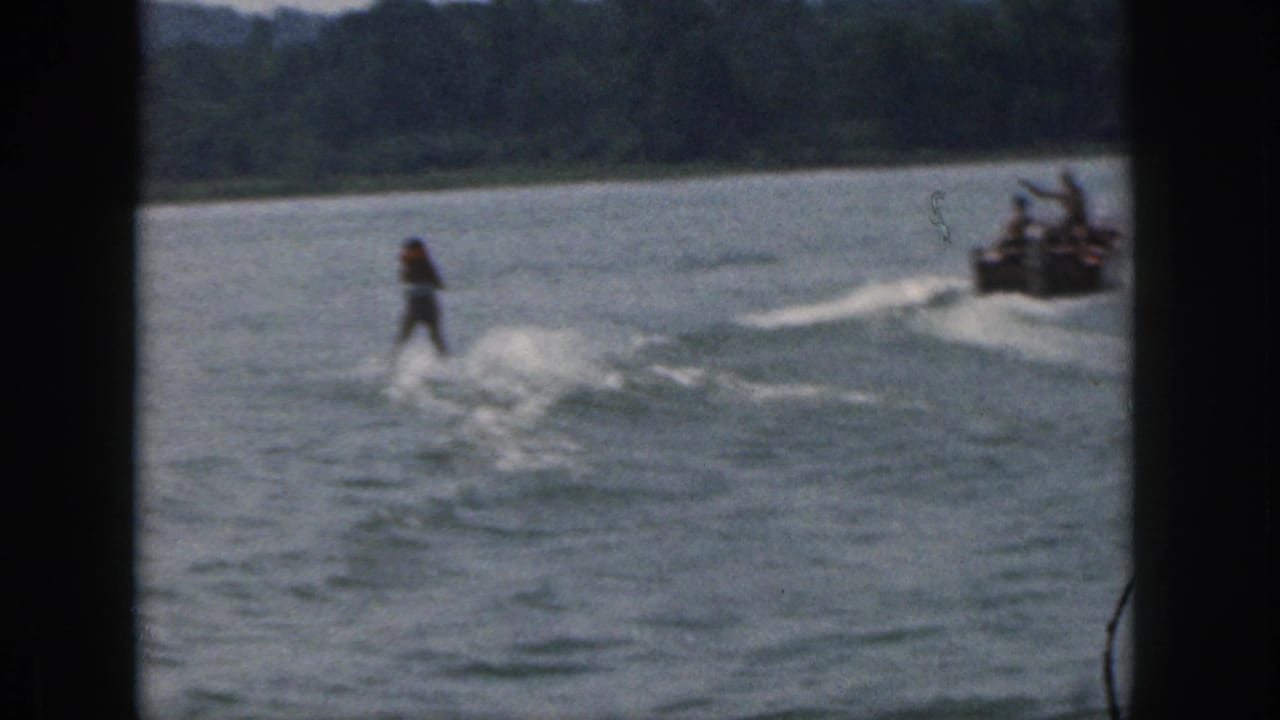 1961 Michigan home-movie frame showing a distant water skier pulled by a speedboat across a wooded lake.