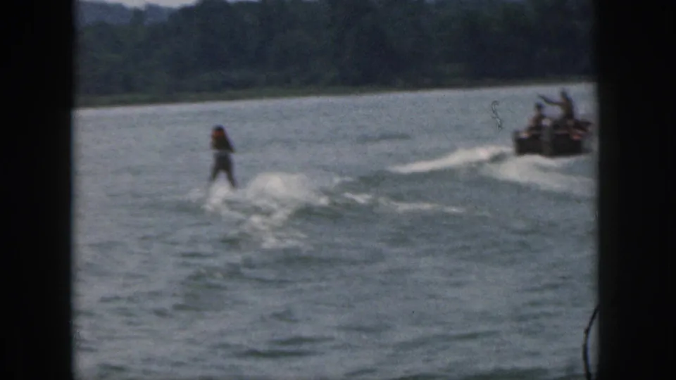 1961 Michigan home-movie frame showing a distant water skier pulled by a speedboat across a wooded lake.