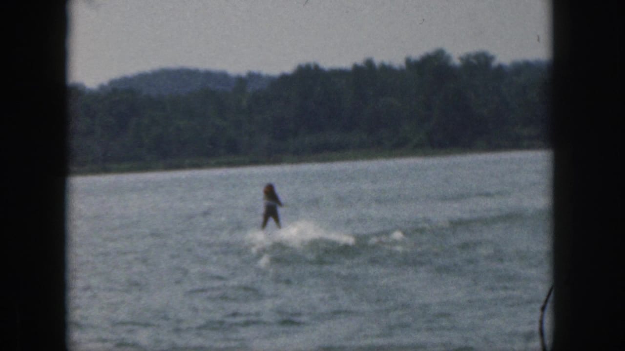 Distant water skier moves across the lake with wake spray and trees along the far shore.