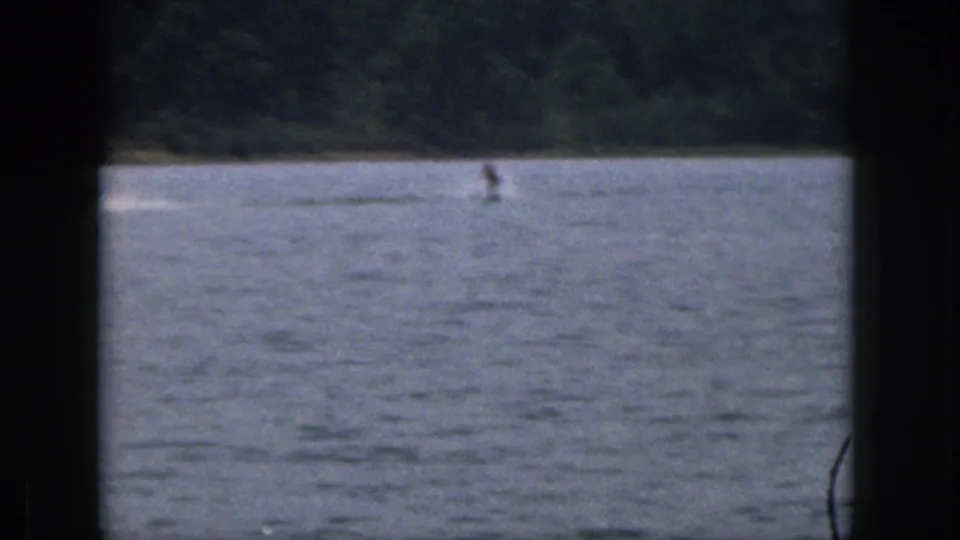 Wide 1961 Michigan lake frame with the water skier small in the distance and the wooded shore behind them.