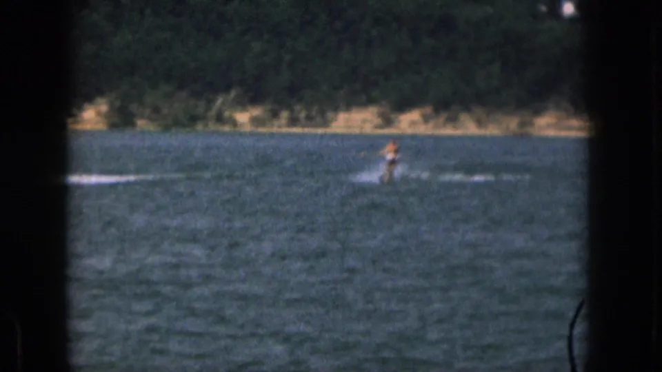 1961 Michigan lake recreation frame with a distant person water skiing across the water near a wooded shore.