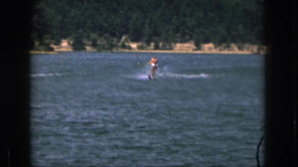 Distant 1961 Michigan home-movie view of a person water skiing on a lake, with spray and trees behind them.