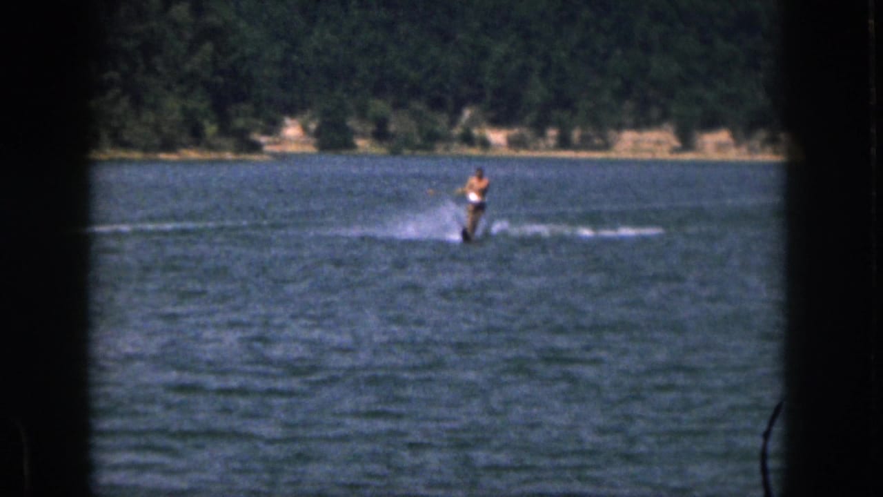 1961 Michigan home-movie frame showing a distant shirtless man water skiing on a lake with trees along the far shore.