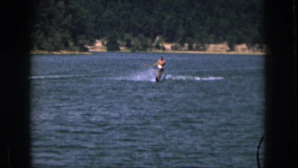 1961 Michigan home-movie frame showing a distant shirtless man water skiing on a lake with trees along the far shore.