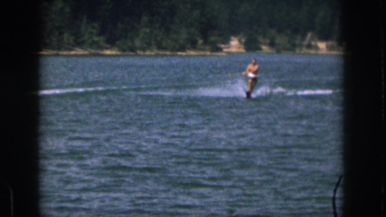 Distant 1961 Michigan lake scene with a shirtless man skiing over rippled water near a tree-lined shore.