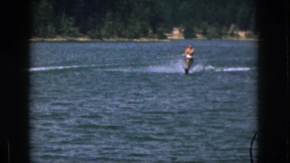 Distant 1961 Michigan lake scene with a shirtless man skiing over rippled water near a tree-lined shore.