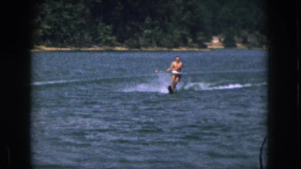 Home-movie frame from Michigan in 1961 showing a shirtless man on a single water ski with a wooded shore behind him.