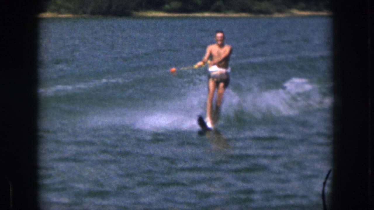 In 1961 Michigan home-movie footage, a shirtless adult man in swim trunks rides water skis across a lake while holding a tow rope with an orange float nearby.